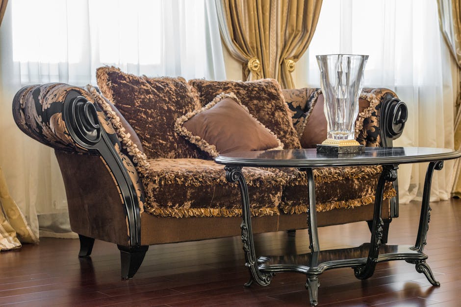 A luxurious living room featuring a vintage-style sofa with plush, patterned upholstery in shades of brown and gold, accompanied by matching cushions. The sofa is positioned near large, floor-to-ceiling windows draped with elegant, gold-toned curtains tied back with decorative tassels. A polished black marble side table with ornate, carved metal legs sits in front of the sofa, holding a tall, clear glass vase with a gold base. The room's hardwood floor is clean and shiny, reflecting the natural light filtering through sheer white curtains behind. This space exemplifies high-end domestic interior with a focus on surface cleaning and maintaining a pristine appearance, reflecting the expertise of Knightsbridge Carpet Cleaners in luxury residential sanitisation and deep cleaning.