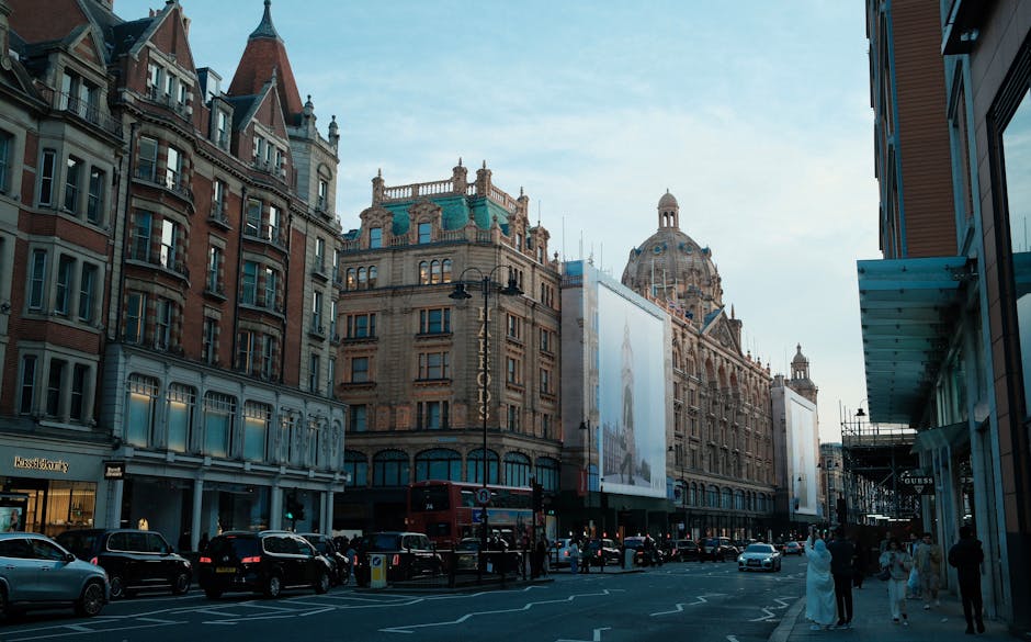 A busy street scene in Knightsbridge, featuring historic and modern buildings with ornate facades and large windows. The street is lined with parked cars and pedestrians walking along the sidewalks. Prominent signage for luxury retail stores is visible, including a high-end jewelry store. The sky above is clear with soft daylight illuminating the buildings, highlighting their detailed architecture and clean windows. The overall scene reflects a well-maintained, upscale commercial area in London. Knightsbridge Carpet Cleaners specializes in surface cleaning, deep cleaning, sanitisation, and domestic cleaning services, ensuring the maintenance of luxury flats and commercial spaces in the area.