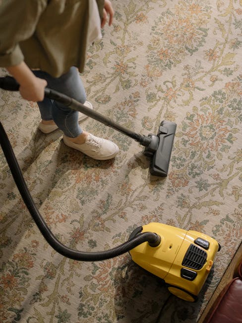A person operating a yellow vacuum cleaner on a traditional patterned area rug, which features floral motifs in beige, green, and orange tones. The individual, dressed in casual clothing including beige shoes and blue jeans, is holding the vacuum's tube and cleaning the carpeted surface within a residential space. The rug appears clean and well-maintained, with clear, vibrant colors and a soft texture. The scene is well-lit, highlighting the detailed design of the carpet. This image showcases surface cleaning and deep cleaning processes commonly associated with domestic cleaning services offered by Knightsbridge Carpet Cleaners, emphasizing professional hygiene and maintenance in a home environment.