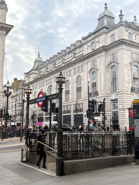Exterior view of Knightsbridge station area showing ornate historic building facades with large windows and detailed stonework, street-level entrance with black iron railings and steps leading down to underground access, surrounded by bustling city traffic and pedestrians, overcast sky providing diffused natural light, with visible signs including the London Underground logo and directional indicators, indicative of an active urban environment typical of central London, captured in daylight. Knightsbridge Carpet Cleaners' service of surface and deep cleaning may target such commercial and residential properties in this area, ensuring hygiene and maintenance of the building surfaces and surroundings.