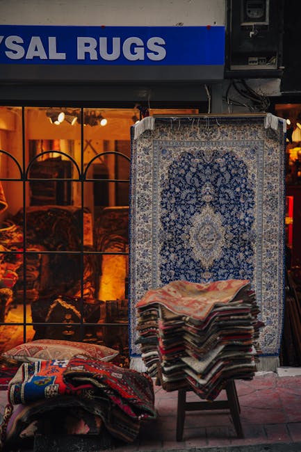 Outside a shop storefront with a sign displaying partially visible text 'AL RUGS', showcasing a display of various rugs. A large, ornate blue and cream patterned rug leans against a stand in front of the shop window, which reveals warm lighting and interior furniture. In front of the shop, several smaller rugs and carpets are stacked on a stool and laid on the pavement, some featuring vibrant, intricate designs. The scene appears clean and well-maintained, with the rugs protected from the elements, highlighting the expertise of Knightsbridge Carpet Cleaners in surface cleaning and maintenance for rugs and carpets. Bright natural light illuminates the display, emphasizing the textures and patterns of the rugs, and the overall image portrays a professional approach to deep cleaning and sanitisation of these textiles for both residential and commercial spaces.