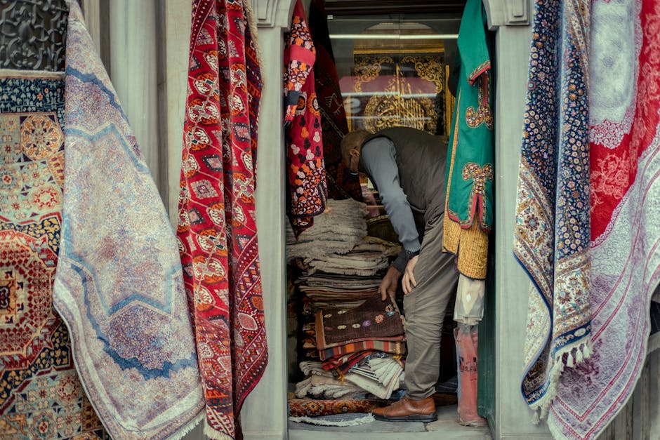 A man examining a high shelf filled with neatly folded woven rugs and carpets inside a shop with an array of colorful rugs hanging on either side. The rugs feature intricate patterns in red, blue, and beige tones, and are displayed on a wall with glossy surfaces. The shop is lit with warm indoor lighting, highlighting the vibrant designs and textures of the textiles. Knightsbridge Carpet Cleaners specializes in surface cleaning and deep cleaning of oriental and decorative rugs, ensuring thorough sanitisation and maintenance to preserve their appearance and longevity. The scene is captured in a commercial setting, emphasizing professional cleaning and routine care for high-value textiles, aligning with Sloane Street Knightsbridge rug cleaning services for shops, Knightsbridge.