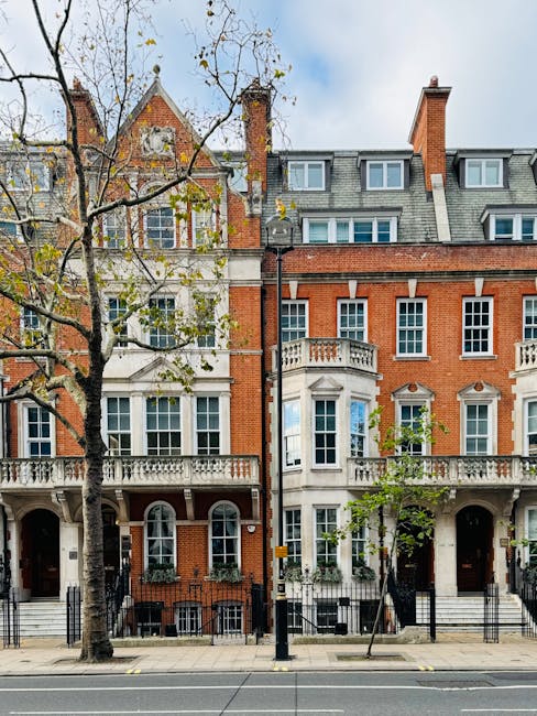 Exterior view of a row of historic red brick and white stone Victorian-style townhouses in Knightsbridge, London, featuring large sash windows, decorative cornices, and small balconies. The sidewalk in front has a row of young trees with sparse leaves, indicative of early autumn, and a black metal fence. The buildings are well-maintained, with clean facades and neat window frames, reflecting a maintained residential area. Bright daylight illuminates the scene, with a partly cloudy sky overhead. External public lighting and black street bollards are also visible, providing a typical urban streetscape. This image exemplifies the classic architectural style prevalent in SW1X Knightsbridge, suitable for discussing surface cleaning, deep cleaning, or preservation in a domestic or commercial property context, as offered by Knightsbridge Carpet Cleaners.