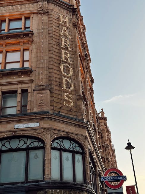Close-up view of the exterior corner of a historic building in Knightsbridge, featuring ornate stonework and large windows with decorative ironwork. The building's facade displays a vertical, illuminated sign spelling 'FEATHERDUSTS,' with string lights draping below. A streetlamp and an underground London Underground sign are visible in the foreground, with a clear sky overhead. The scene captures the classic architectural style typical of the area, emphasizing detailed craftsmanship and urban cleanliness, relevant to comprehensive surface cleaning and maintenance services offered by Knightsbridge Carpet Cleaners for domestic and commercial properties.