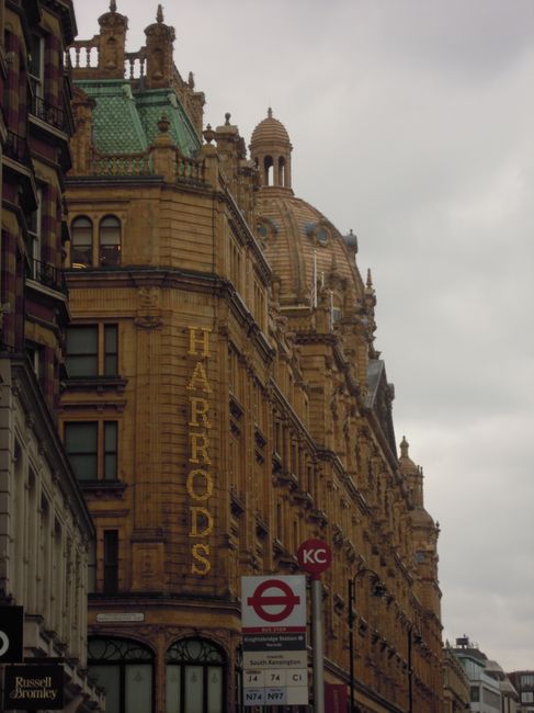 A close-up view of a historic sandstone building in Knightsbridge, featuring ornate architectural details, dome-shaped towers, and intricate carvings on the facade under an overcast sky. The building has large windows with decorative framing, and a vertical sign reading 'Harrods' is visible on the corner. In the foreground, there is a London Underground station entrance with a red circle and a blue bar, displaying tube line information. The scene captures the grandeur and architectural elegance typical of the area, emphasizing the urban environment and classic design elements, with natural diffused lighting and clean surfaces. Knightsbridge Carpet Cleaners would recommend regular surface cleaning to maintain the building's appearance and hygiene.