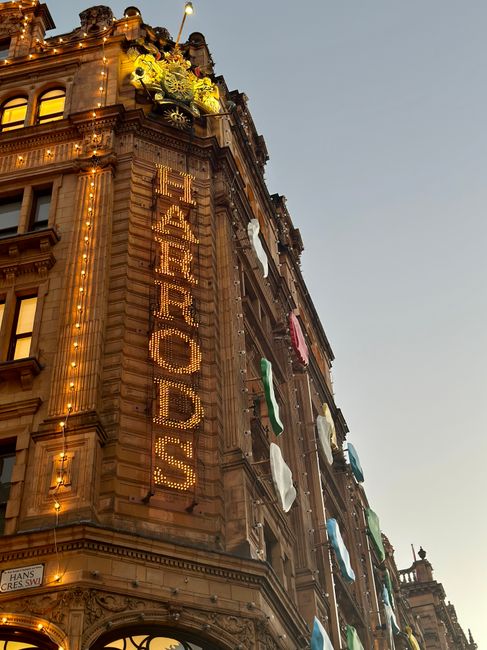 A close-up view of a historic sandstone building in Knightsbridge, featuring ornate architectural details, dome-shaped towers, and intricate carvings on the facade under an overcast sky. The building has large windows with decorative framing, and a vertical sign reading 'Harrods' is visible on the corner. In the foreground, there is a London Underground station entrance with a red circle and a blue bar, displaying tube line information. The scene captures the grandeur and architectural elegance typical of the area, emphasizing the urban environment and classic design elements, with natural diffused lighting and clean surfaces. Knightsbridge Carpet Cleaners would recommend regular surface cleaning to maintain the building's appearance and hygiene.